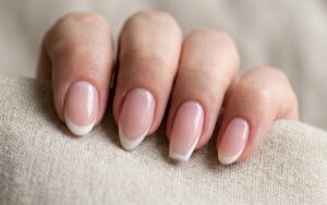 Close-up of a hand with natural French manicure nail shapes resting on a textured beige fabric surface, showcasing a clean and elegant nail design.