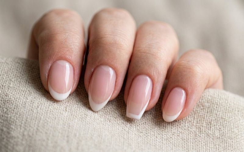Close-up of a hand with natural French manicure nail shapes resting on a textured beige fabric surface, showcasing a clean and elegant nail design.